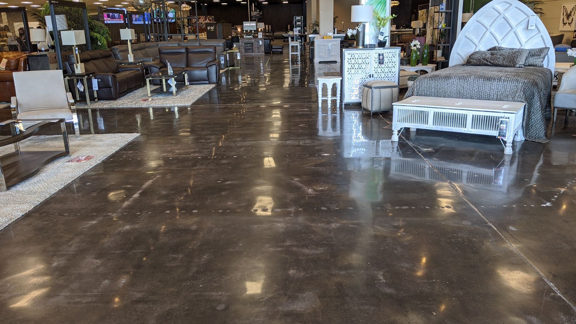 Polished, dark floor of a furniture showroom reflects displays, including a bed and sofas.