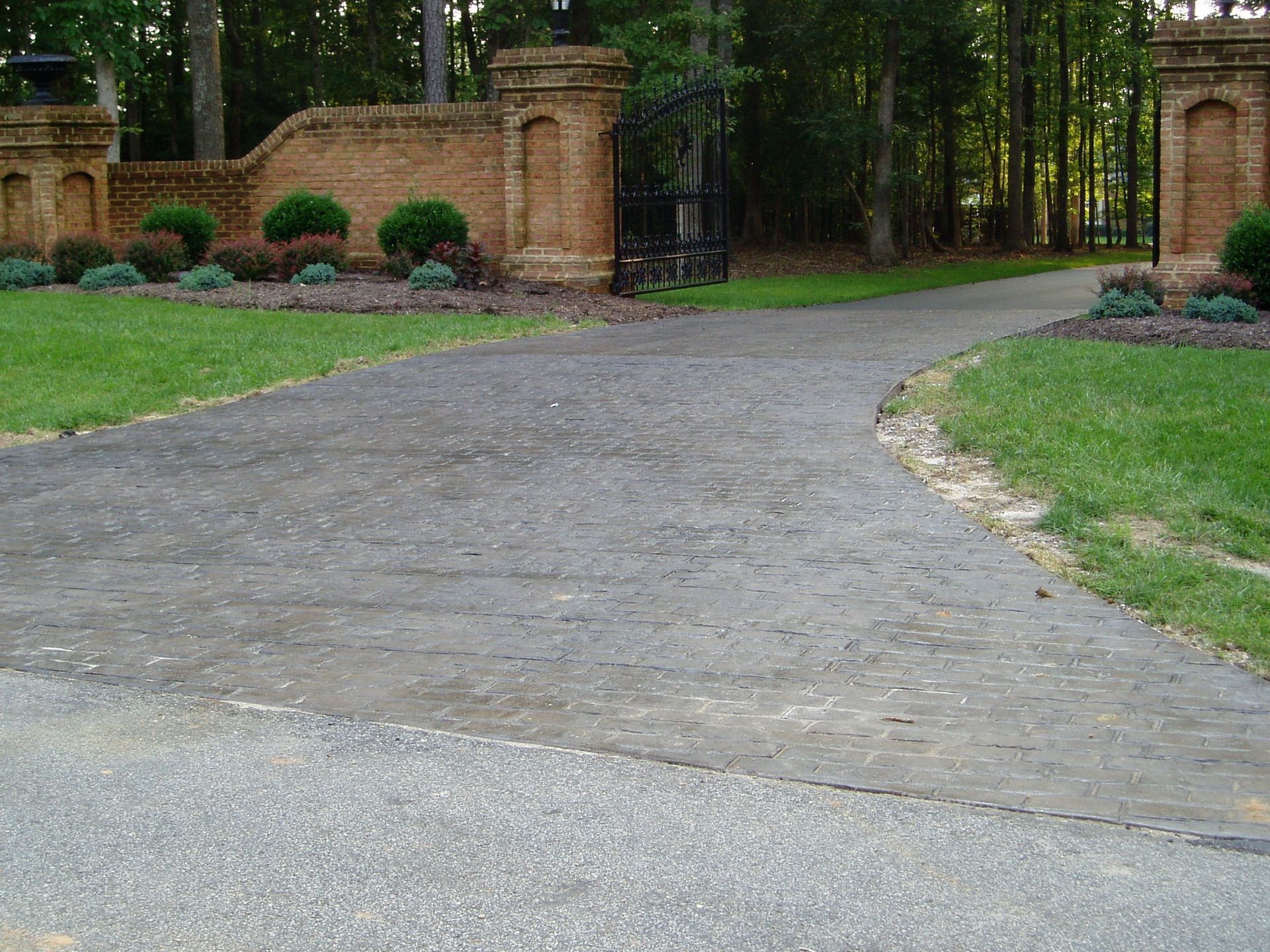 Gravel driveway curves to a brick gate, flanked by green lawns and shrubs. Forest in background.