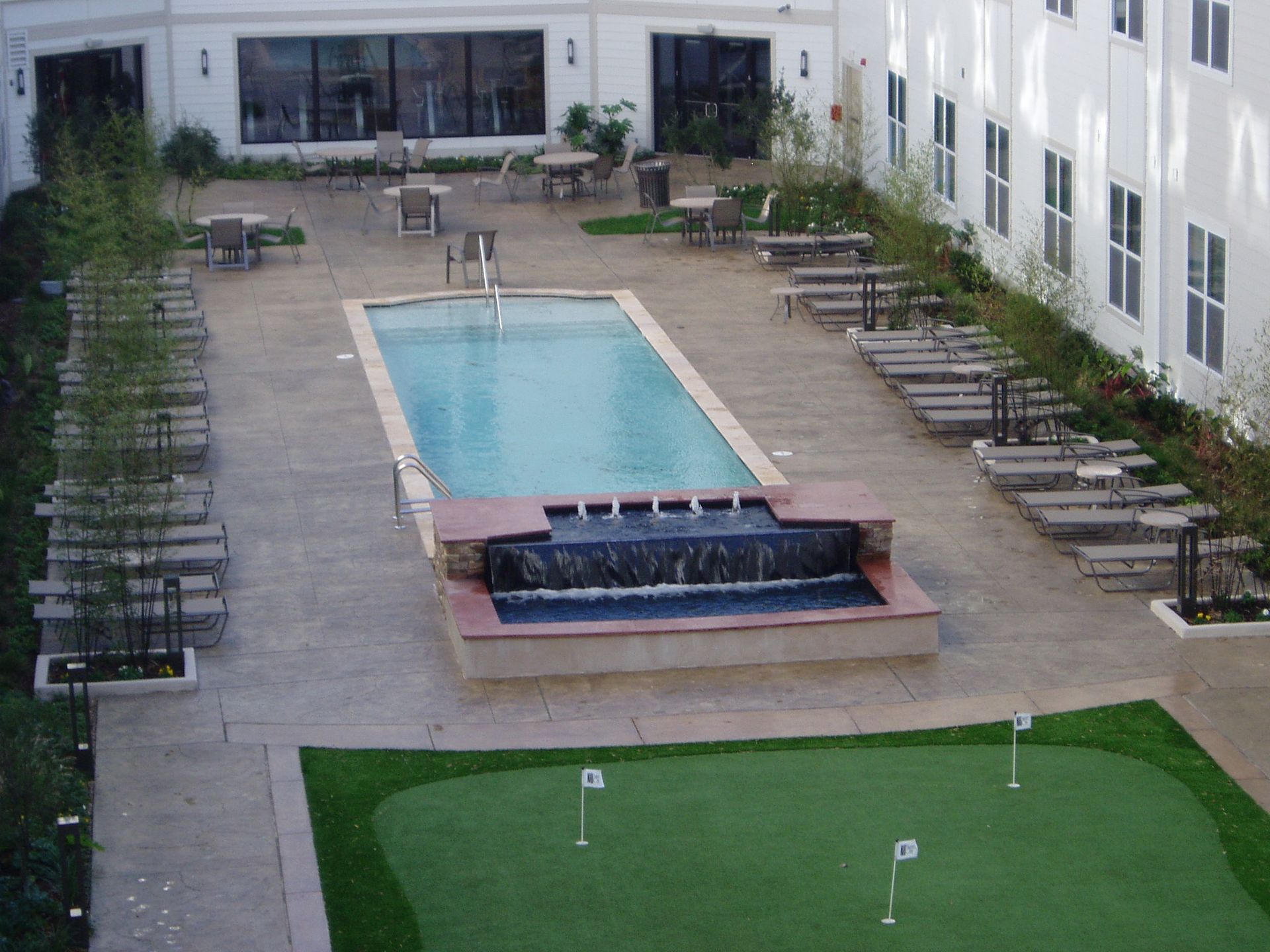 Courtyard with rectangular pool, waterfall, putting green, lounge chairs, tables, and building.