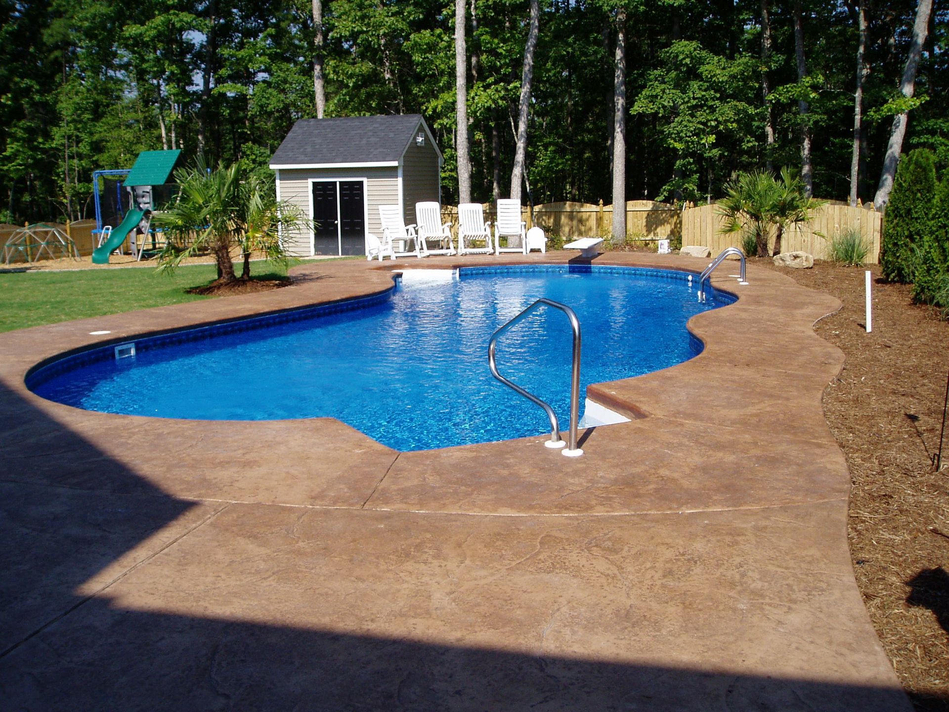 A blue, kidney-shaped swimming pool with a brown concrete patio, shed, and surrounding trees.