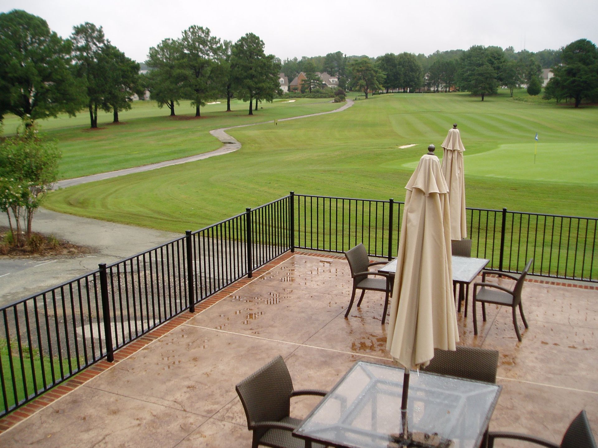 Outdoor patio overlooking a golf course. Tables, chairs, and umbrellas are on the patio. Green grass, trees, and a path are visible.