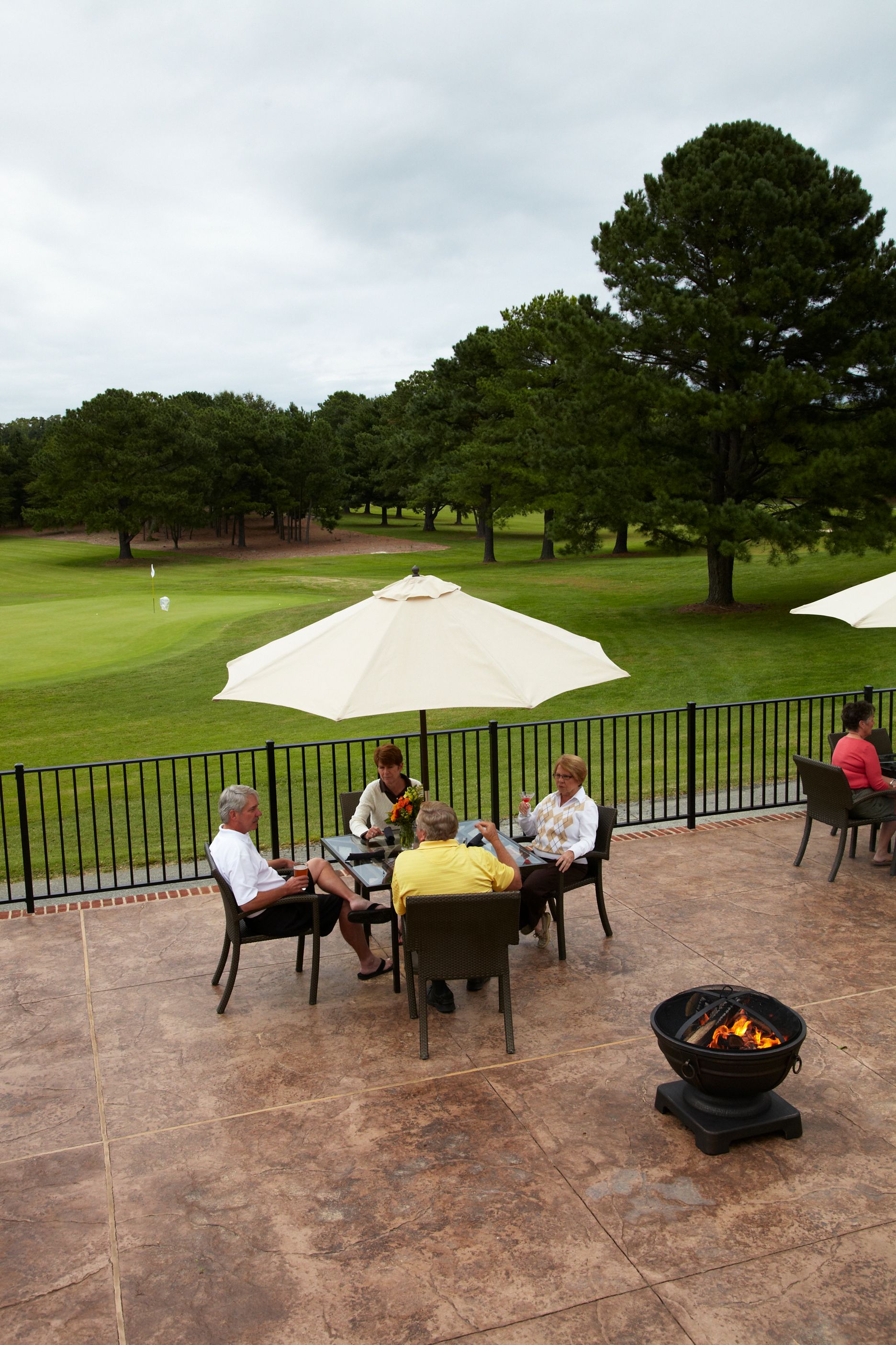 People sit at a table on a patio overlooking a golf course, beneath a cream-colored umbrella.
