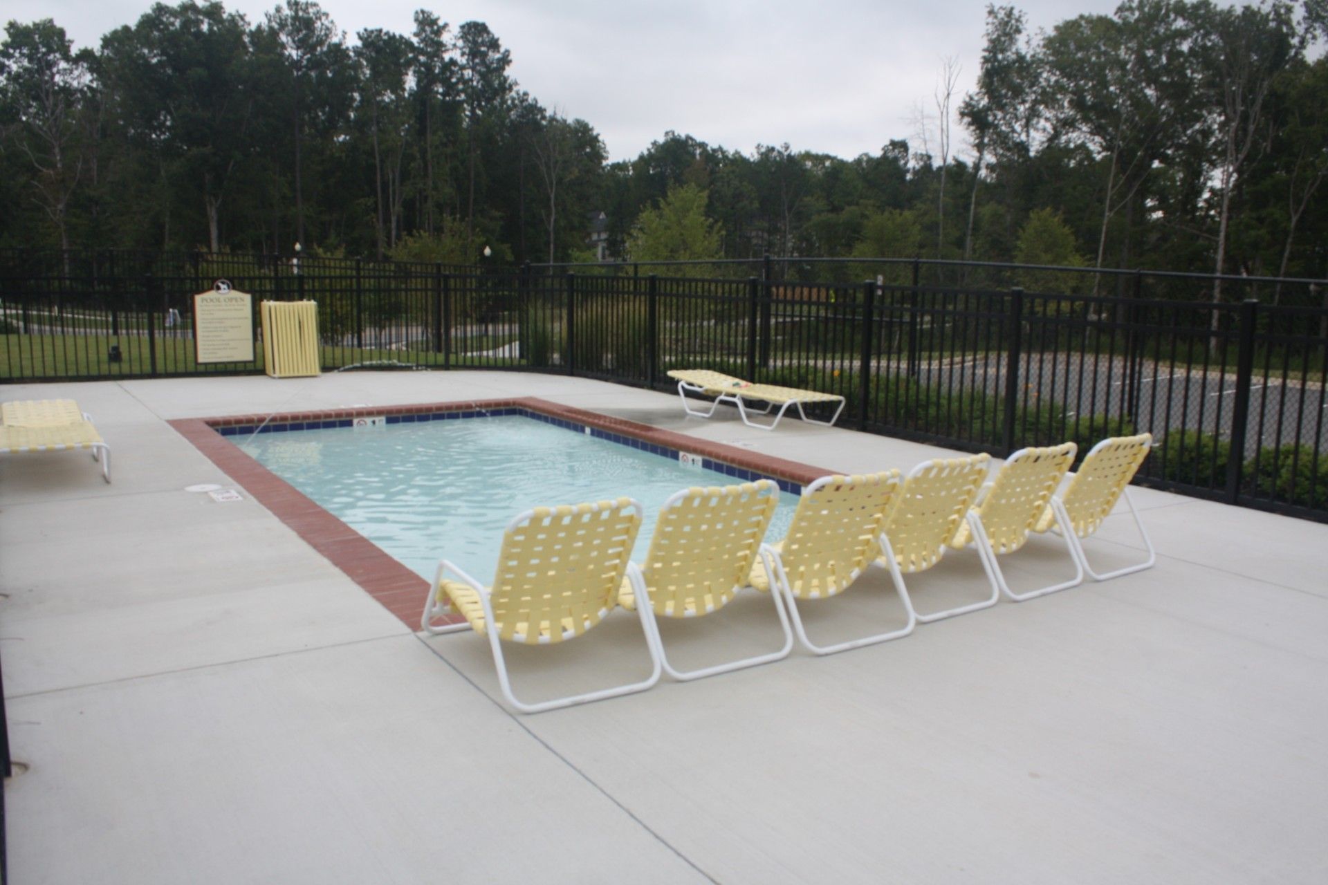 Pool with yellow lounge chairs on a concrete patio, surrounded by a black fence and trees.