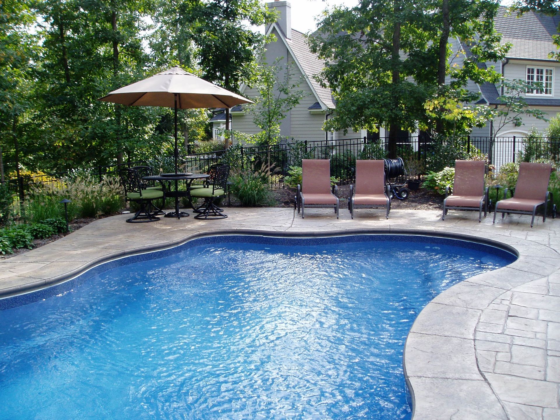 Swimming pool with lounge chairs, umbrella, and table on a patio. Green foliage and a house in the background.