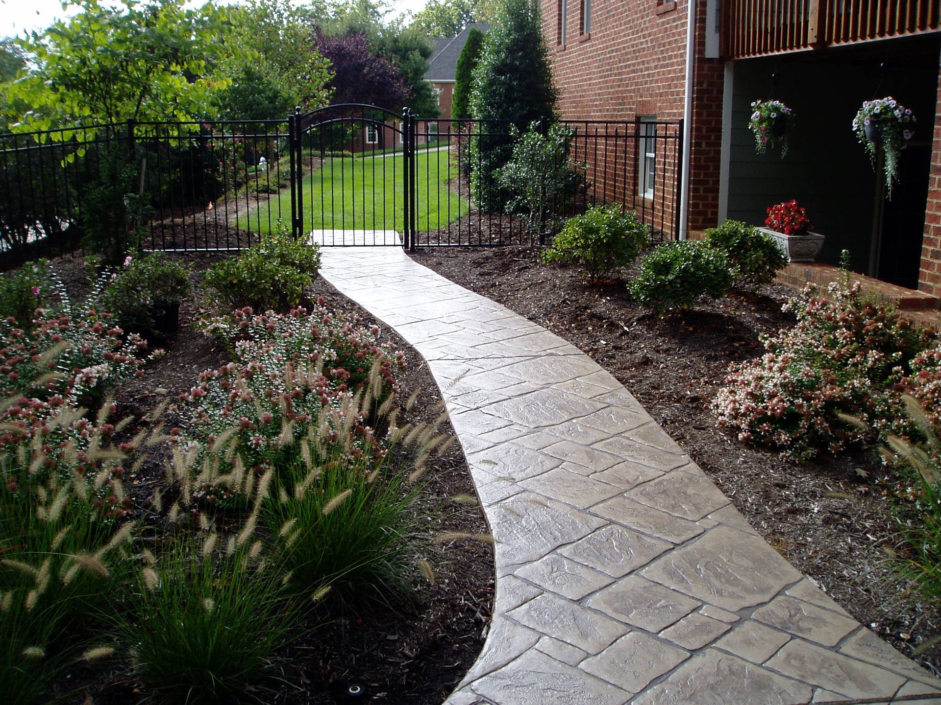 Winding stone pathway through a garden, leading to a black gate. Lush green plants and brown mulch on either side.