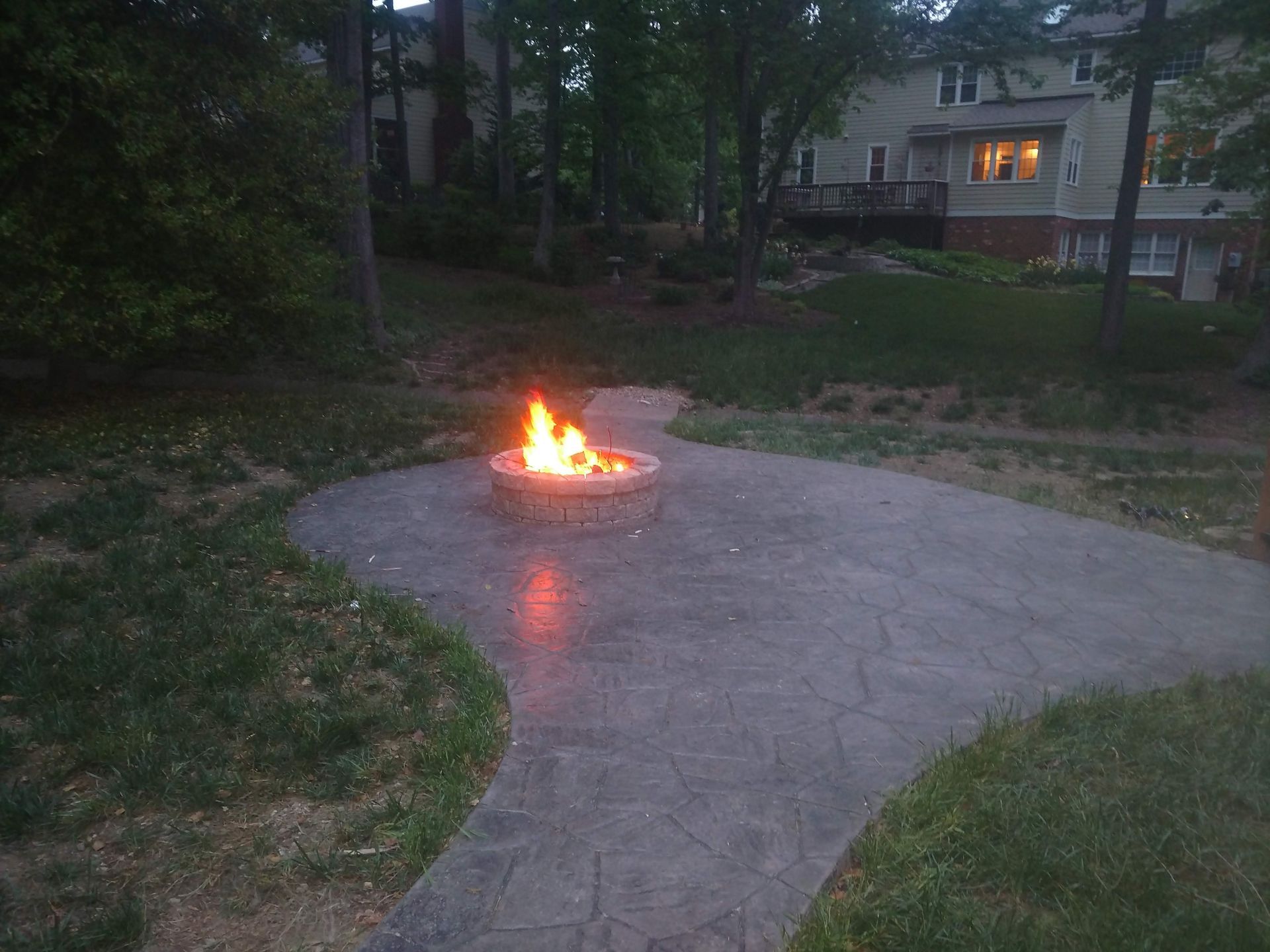 Fire burning in a stone fire pit on a curved patio, surrounded by grass and trees, a house in the background.