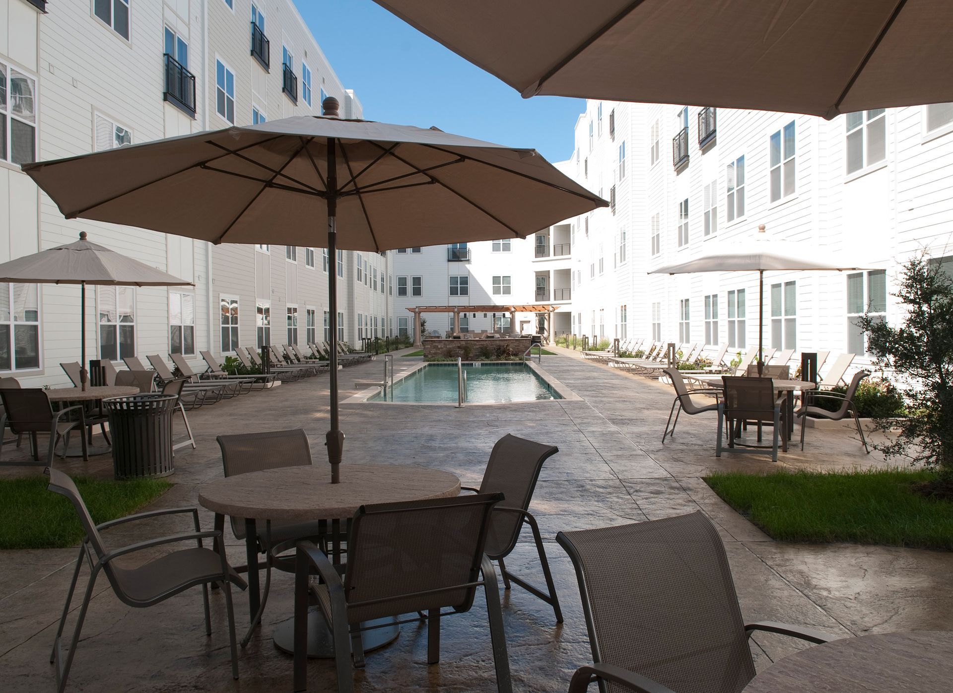Courtyard with tables, chairs, umbrellas, and pool surrounded by multi-story white buildings on a sunny day.