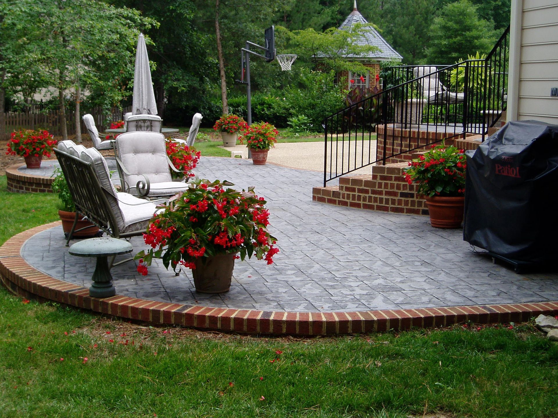 Brick-lined patio with seating, surrounded by potted red flowers, leading to a garden with a gazebo.