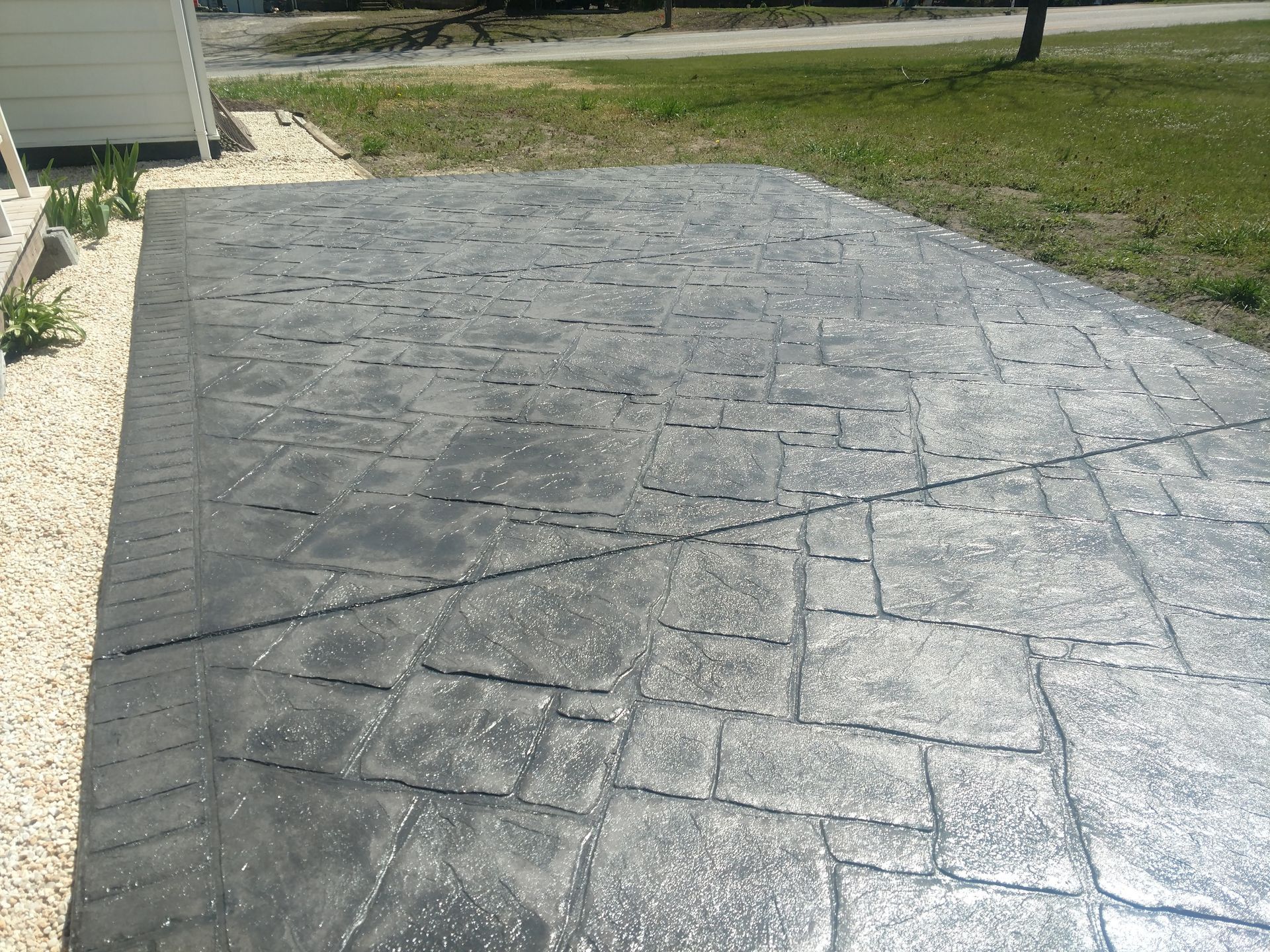 Stamped concrete patio with a textured, stone-like pattern; gray color. Gravel border, grass, and a sunny outdoor setting.