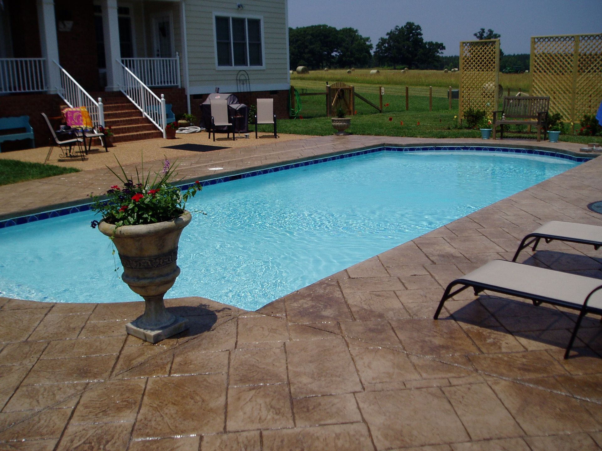 Pool with blue water and surrounding stone patio, next to a house with a lawn.