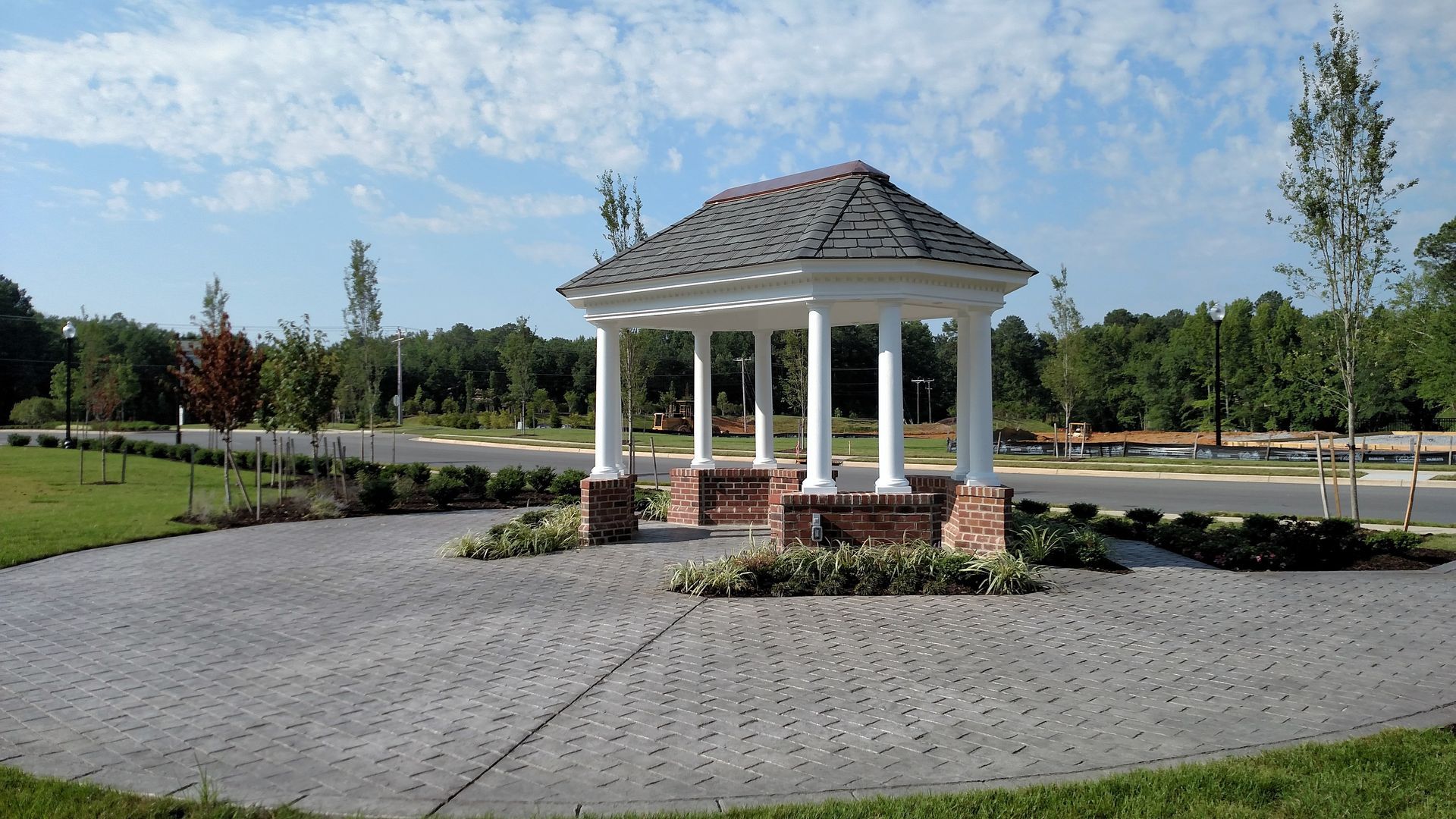 Gazebo with white columns and brick base in a paved area, with trees and blue sky.