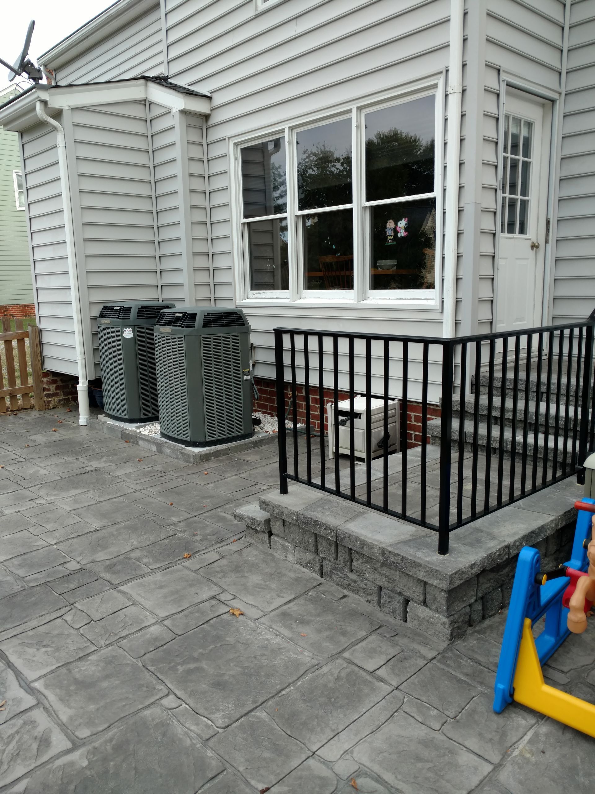 Backyard patio with AC units next to a house with a raised stone step and black railing.
