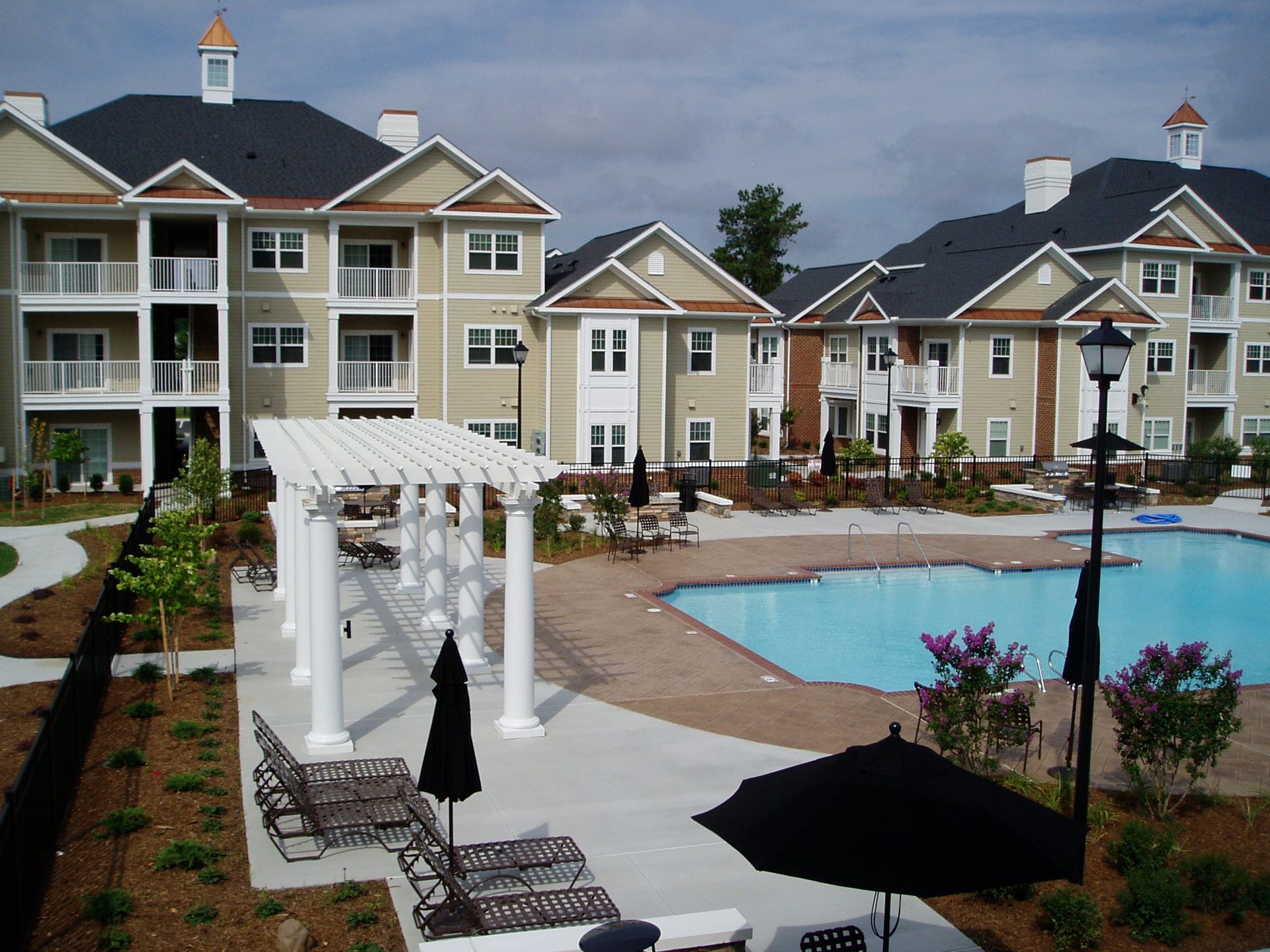 Apartment complex with a swimming pool and pergola. Beige buildings with dark roofs.