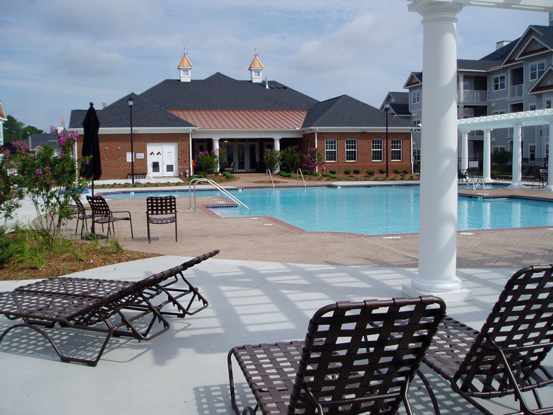 Swimming pool with lounge chairs, building with a red brick facade in the background. Sunny day.