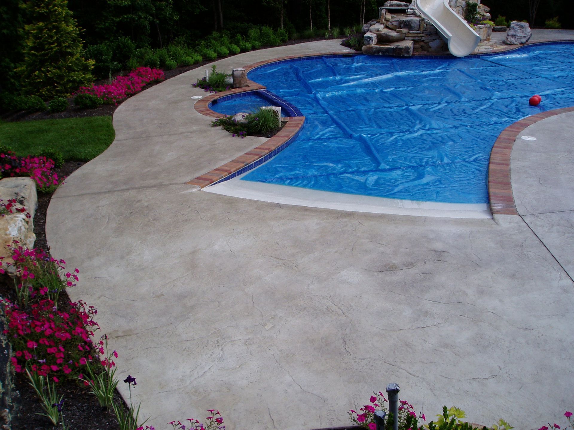 Pool with blue cover, surrounded by concrete patio and flowerbeds; a hot tub is nearby.