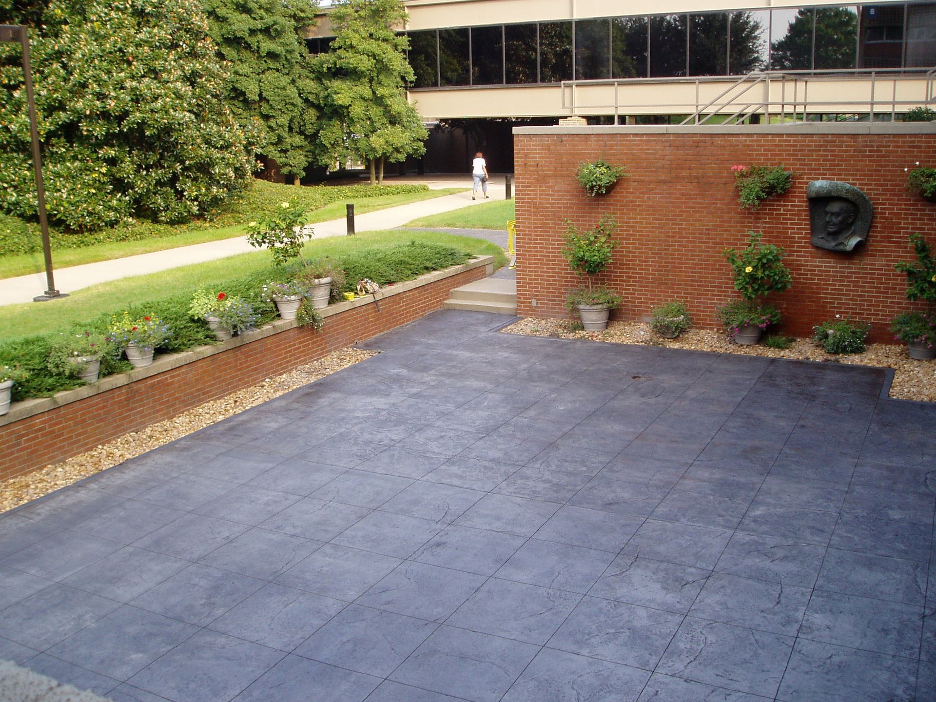 Brick patio with gray tiles, surrounded by a brick wall with potted plants and landscaping in front of a building.