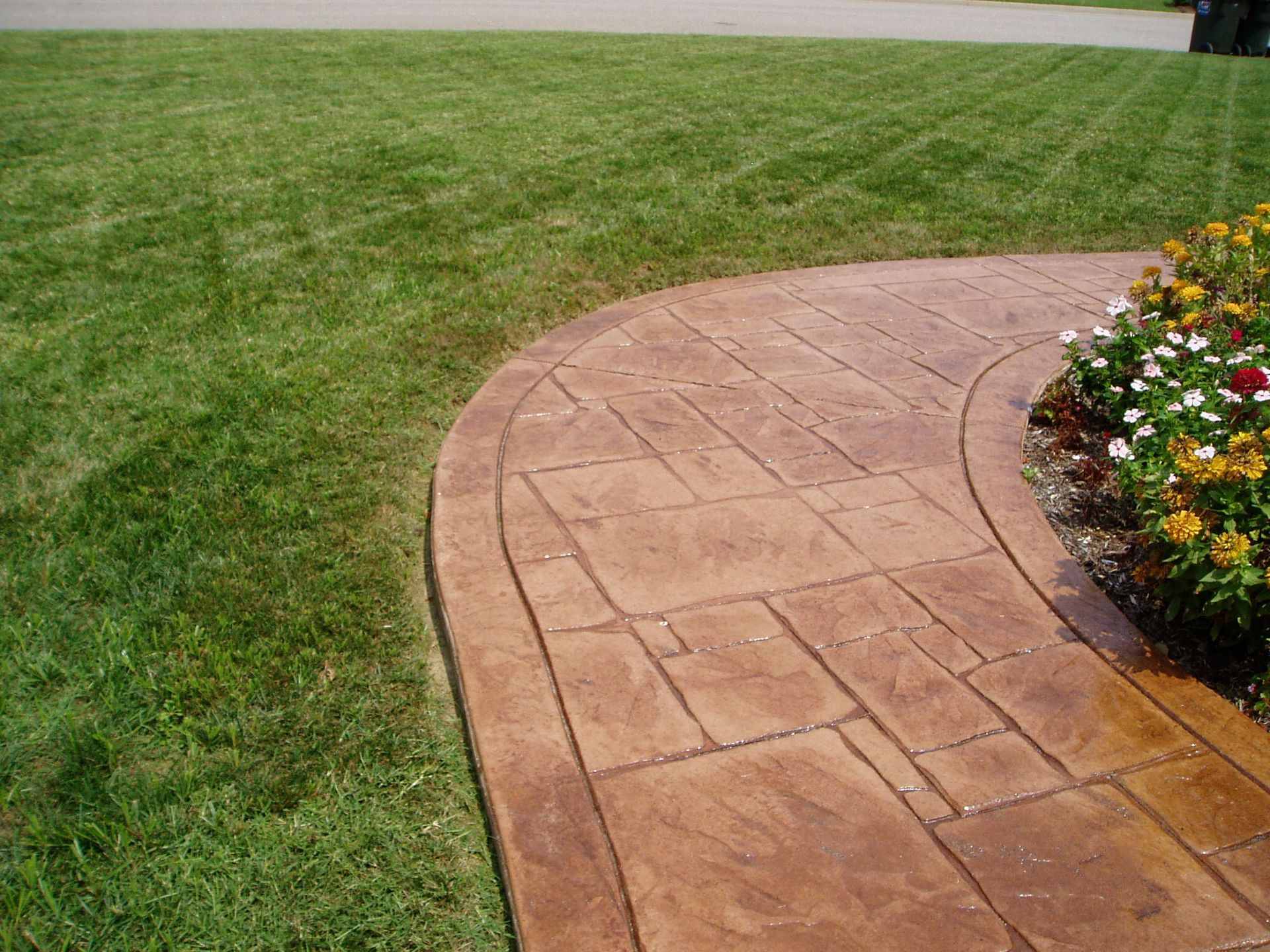 Brown stamped concrete pathway curves through green lawn, next to flower bed with yellow and red flowers.