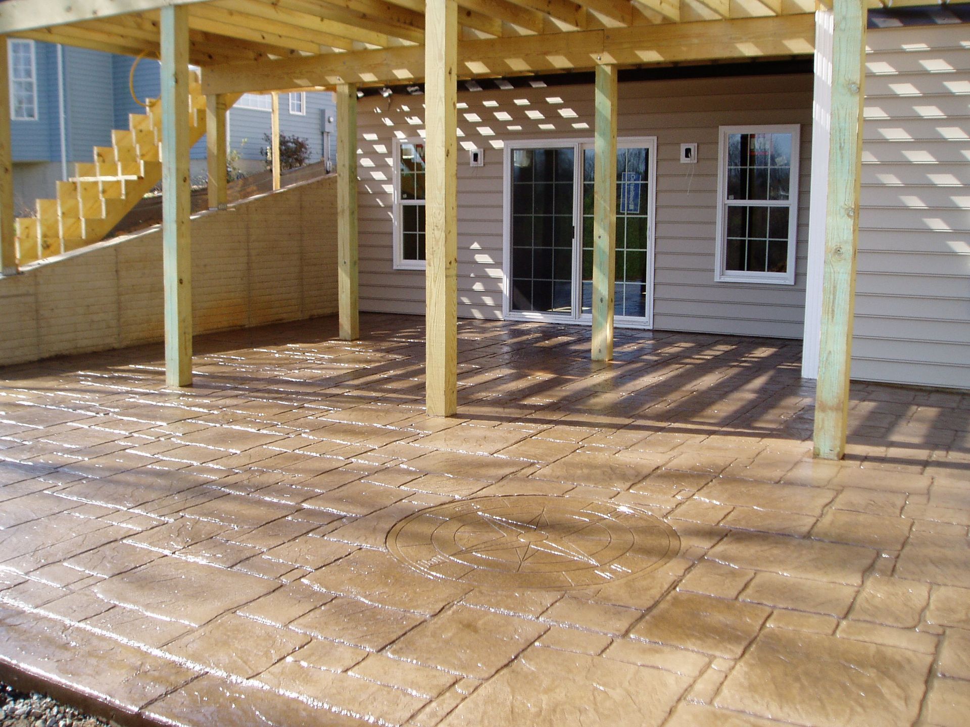Stamped concrete patio under a wooden deck with stairs. House exterior visible, sunlight and shadows.