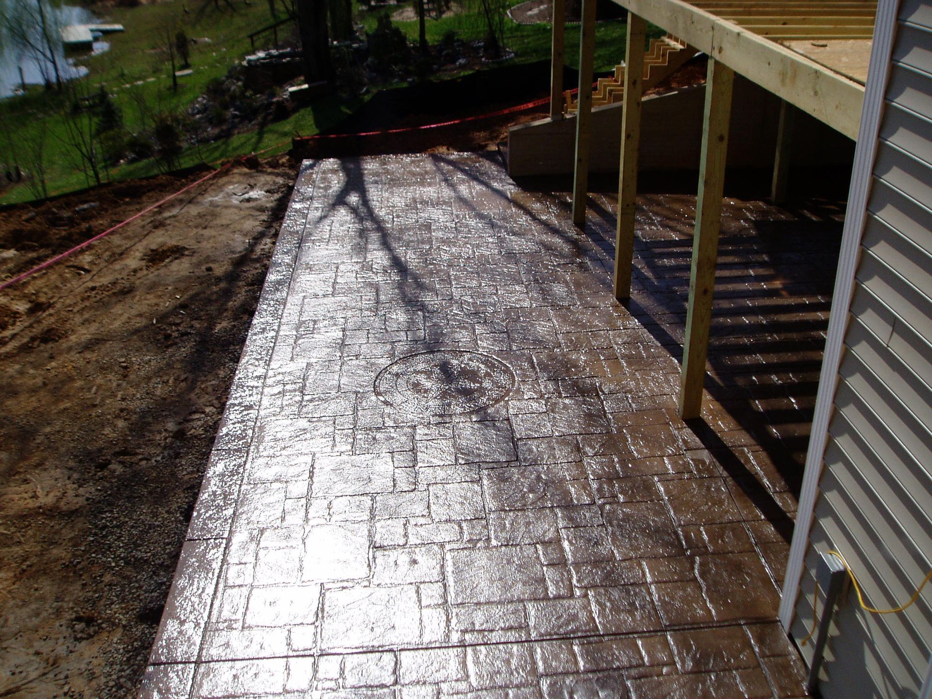 Stained, stamped concrete patio and walkway next to a house, leading to a wooden deck with a lake visible in the background.