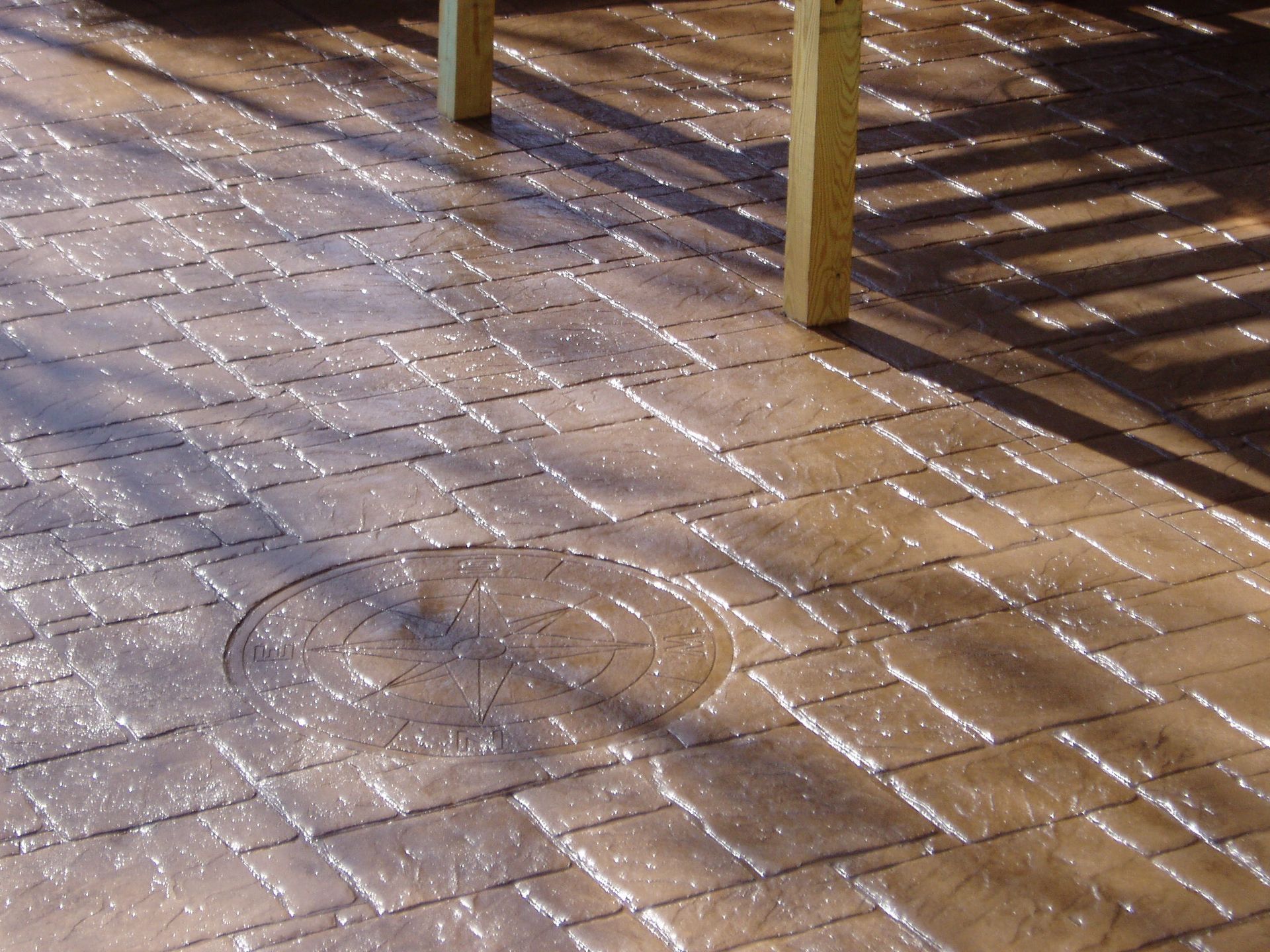 Stamped concrete patio with a brick pattern, brown with a circular design, and wooden support beams casting shadows.
