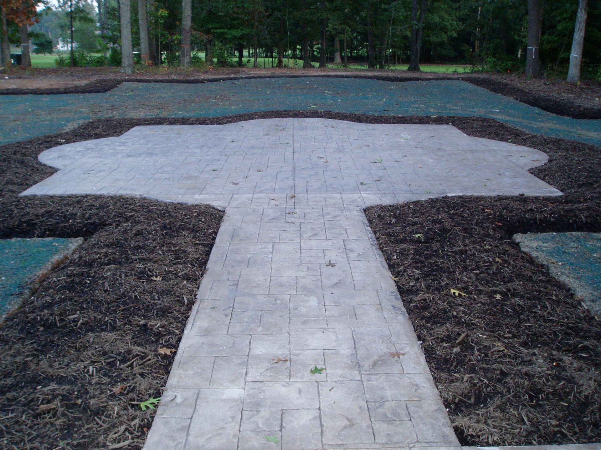 Concrete patio and walkway with dark mulch border, surrounded by green turf. Trees in the background.
