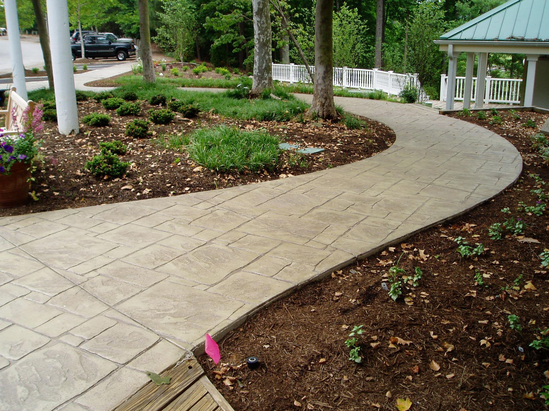 A curved concrete walkway through a landscaped garden with trees, plants, and a gazebo.