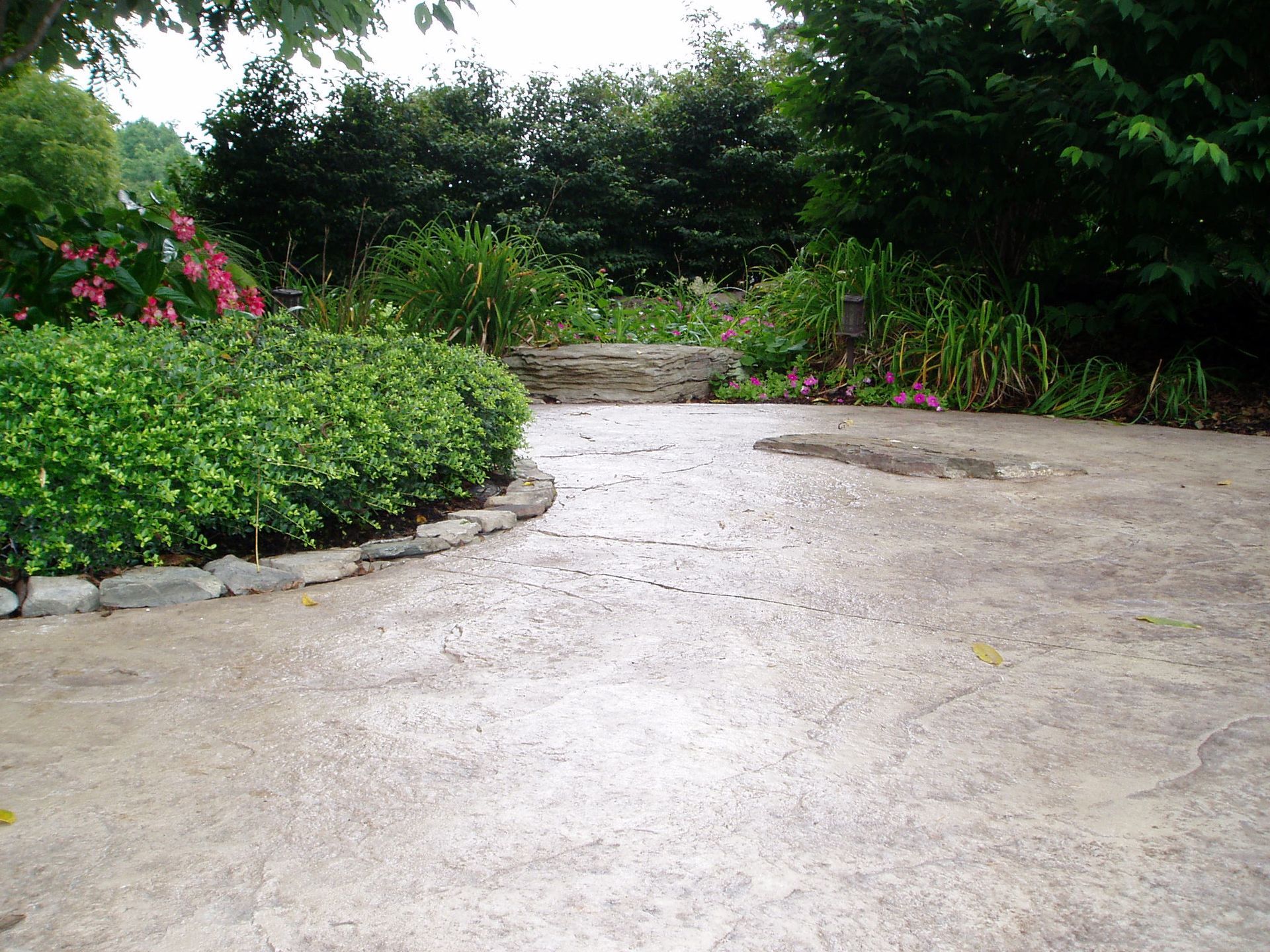 Concrete path curves through a garden with green hedges and colorful flowers.