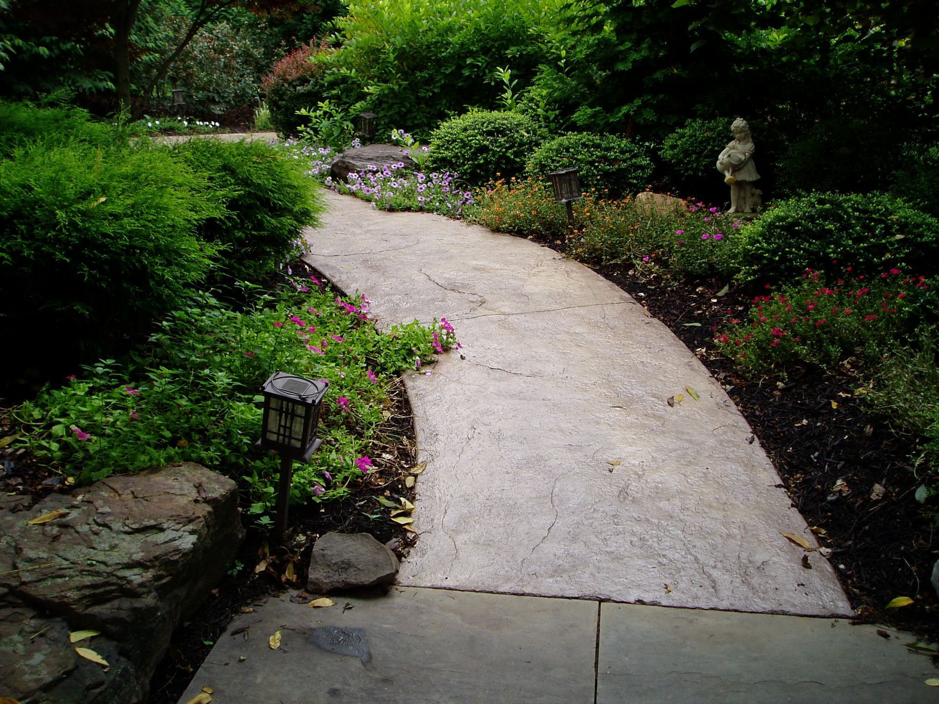 Concrete path winds through a garden with lush greenery and small solar lights.