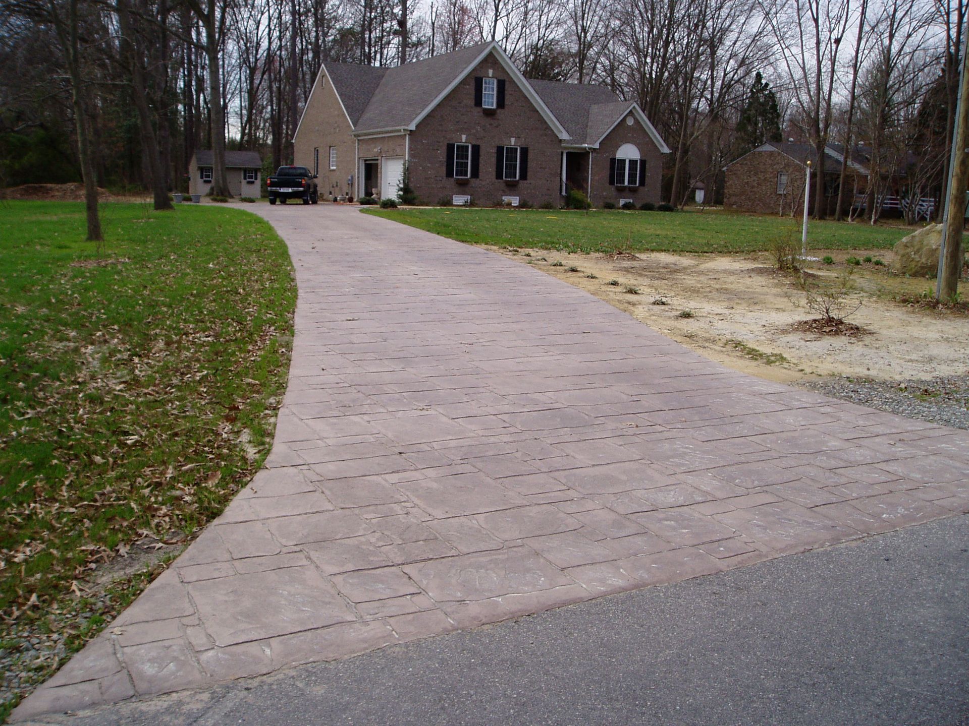 Brick paver driveway leading to a brick house with black shutters, trees, and grass.
