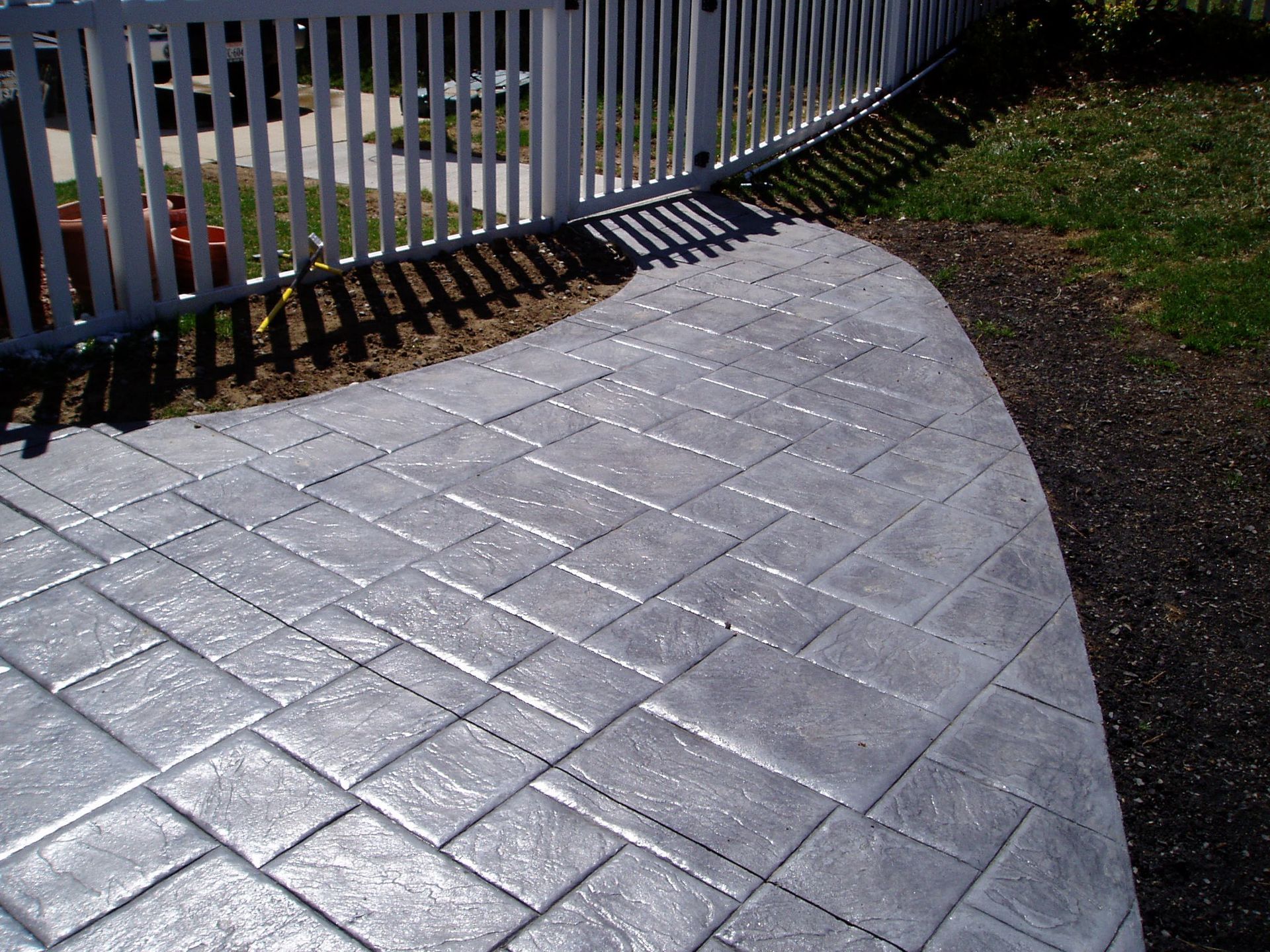 Stamped concrete walkway with a brick pattern next to a white picket fence and green grass.