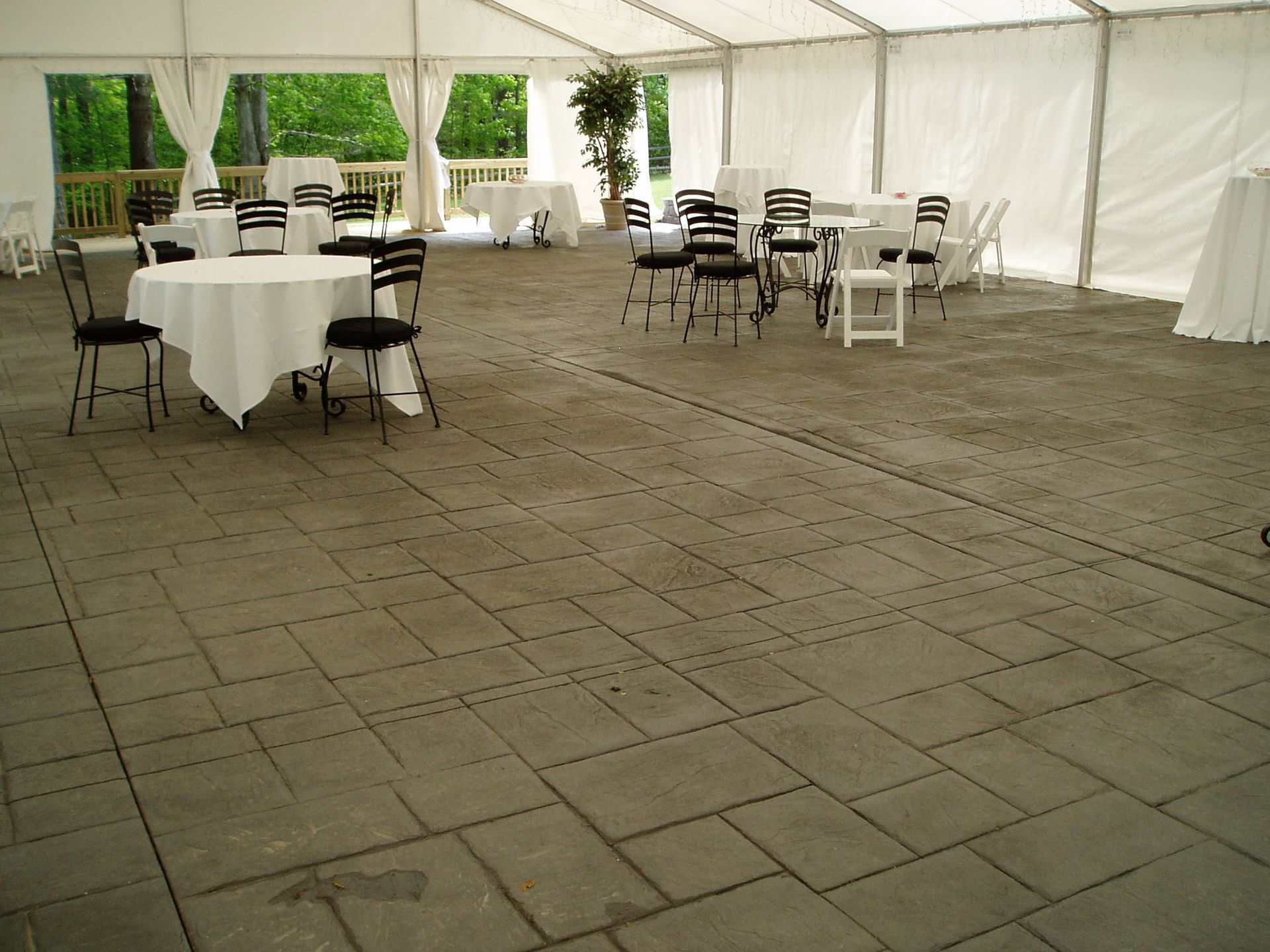 Inside a white tent: tables with white tablecloths, black chairs on a gray stone floor, and a forest backdrop.
