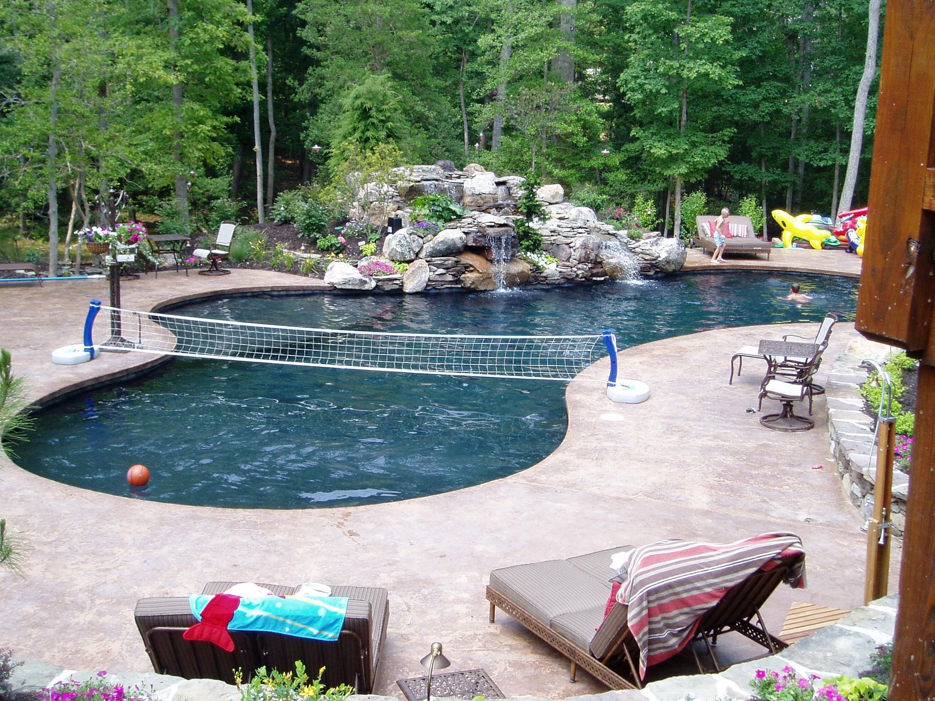 Swimming pool with waterfall feature, volleyball net, and lounge chairs surrounded by trees.