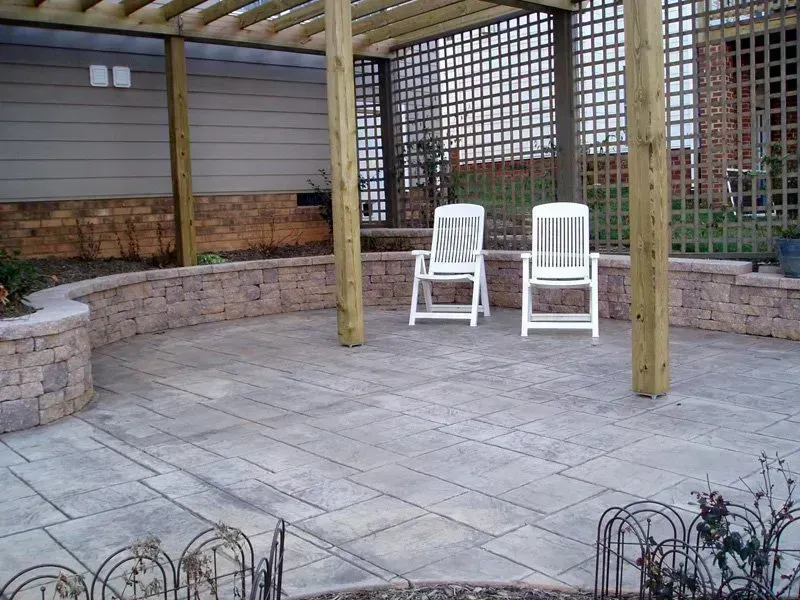 Stone patio with a pergola, two white chairs, and a curved brick wall.