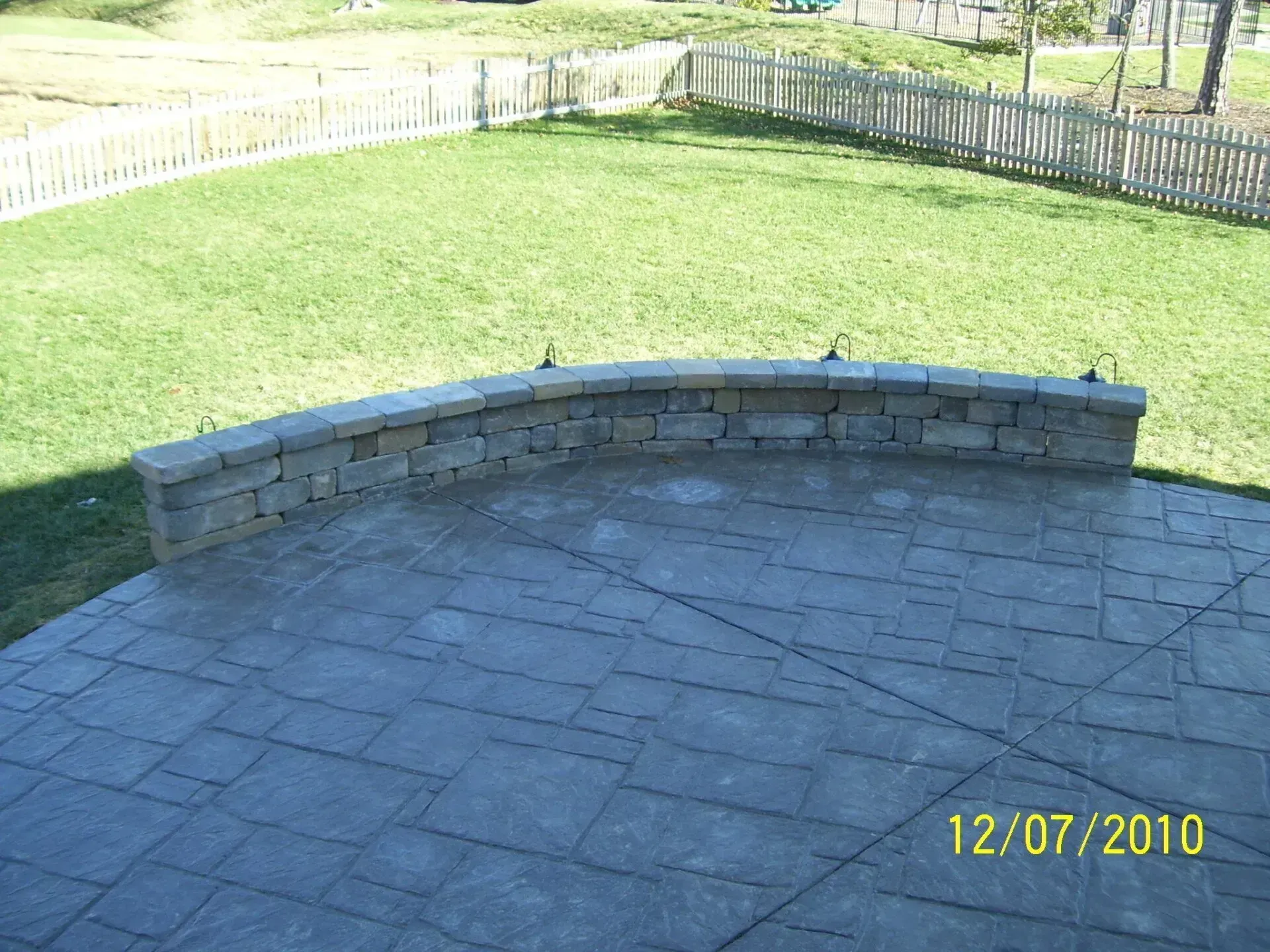 Curved stone retaining wall on a concrete patio with grass and a fence in the background.