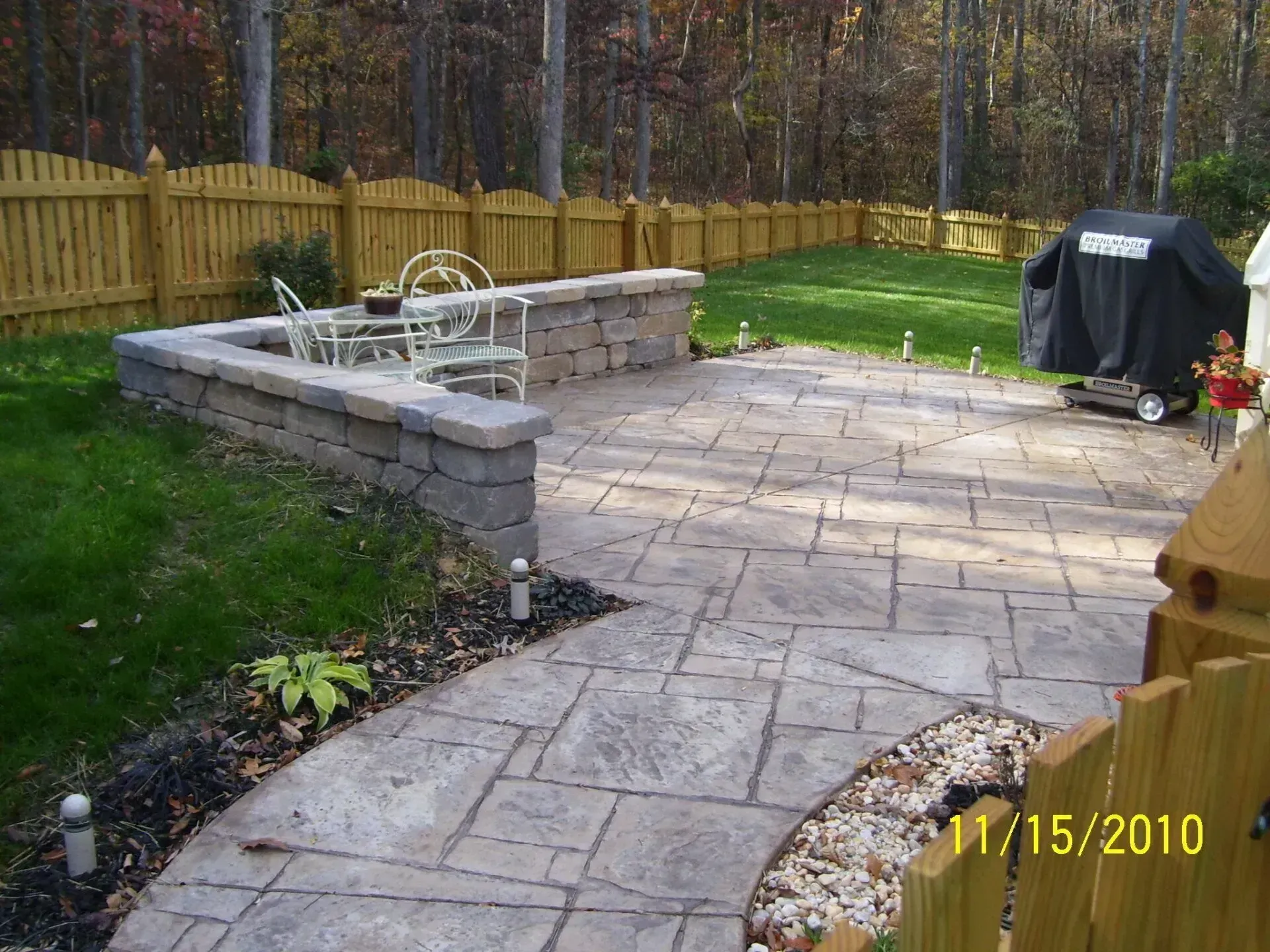 Patio with stone pavers, retaining wall, and fenced backyard with a grill.