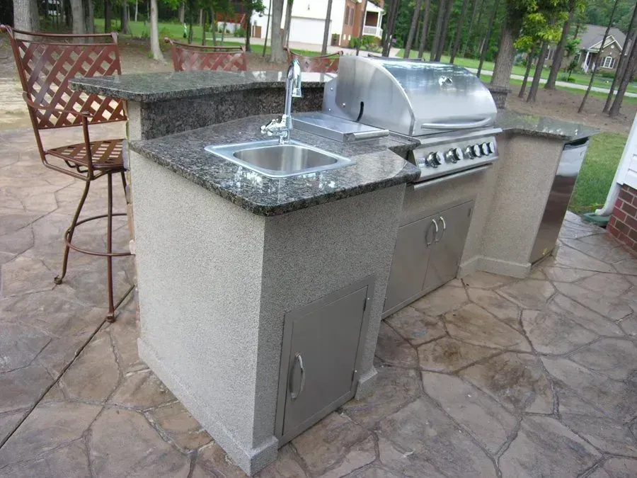 Outdoor kitchen with a grill, sink, and cabinets on a stone patio, with a bar stool.