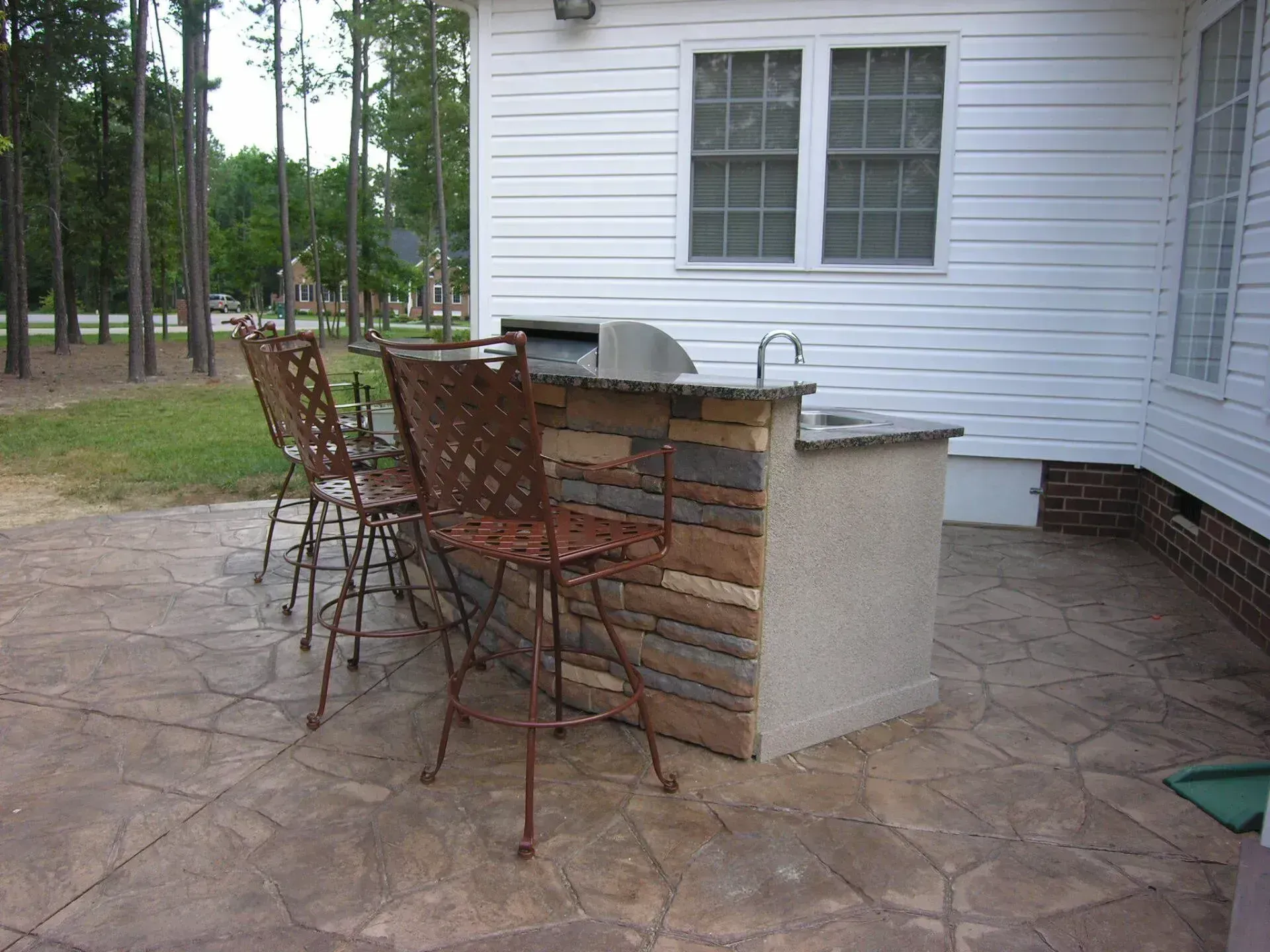 Outdoor kitchen with grill, sink, and bar seating on a stone patio next to a white house.