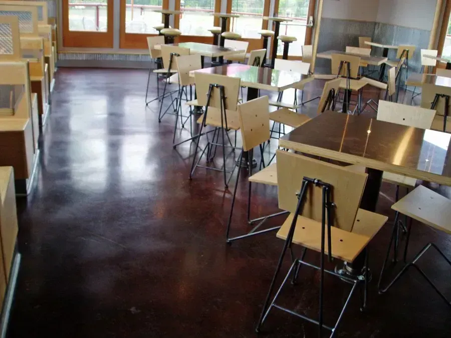 Cafeteria with light wood tables and chairs, dark stained concrete floor, and large windows.