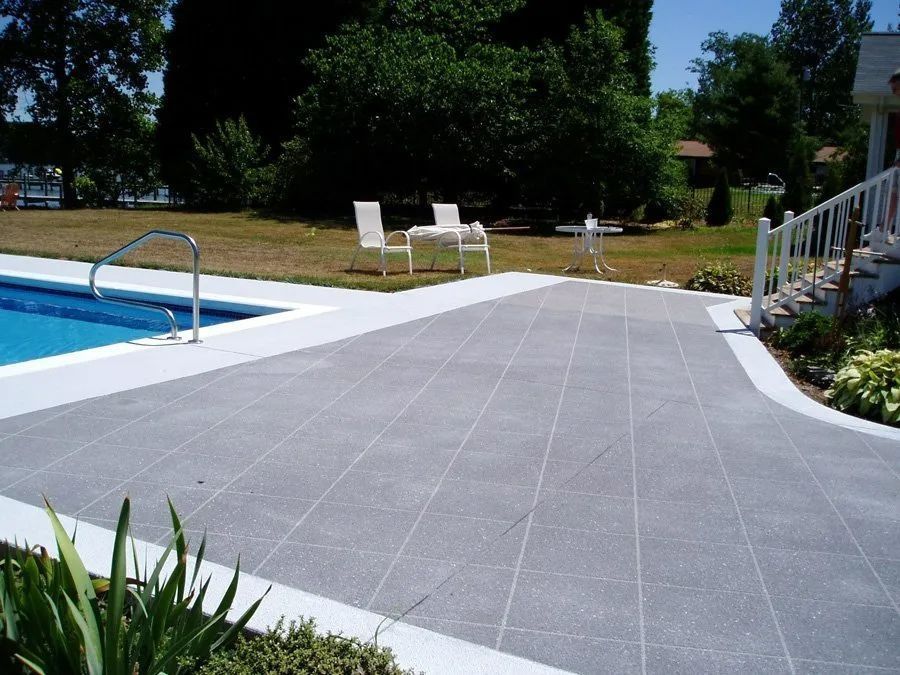 Poolside patio with gray tiled surface and white coping; two white chairs, table.
