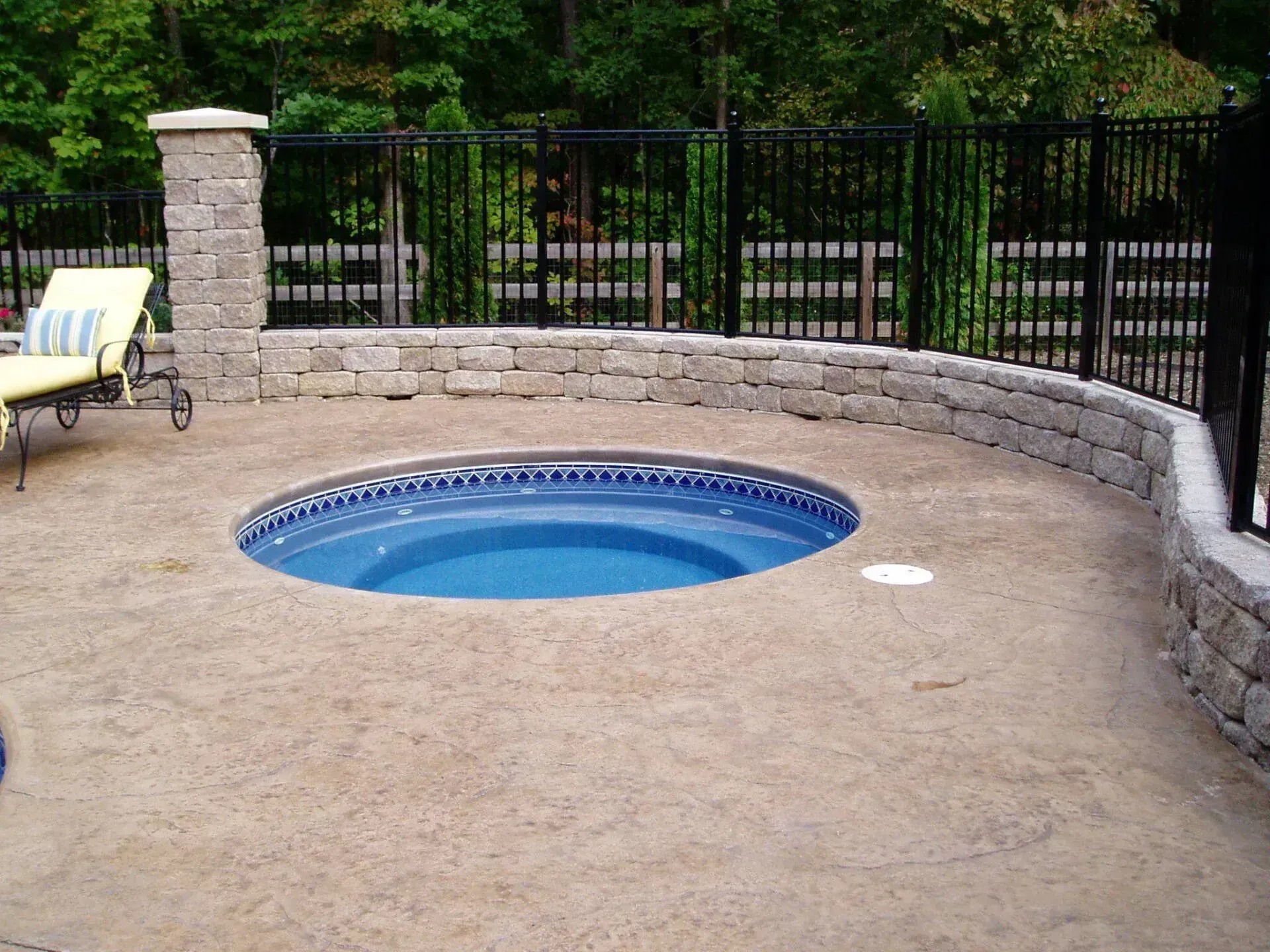 Hot tub in a concrete patio surrounded by a low stone wall and black metal fence.