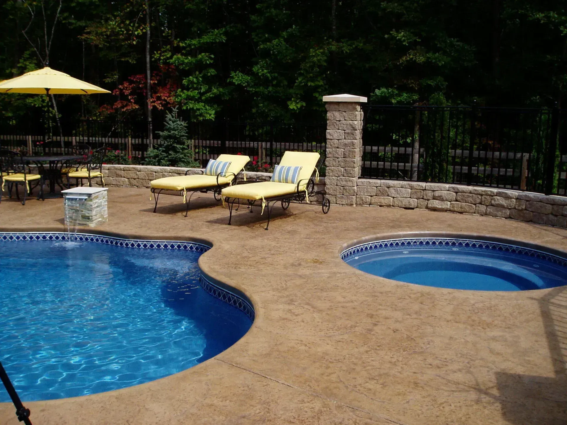 Pool and spa with lounge chairs and umbrella on a patio surrounded by stone walls and trees.