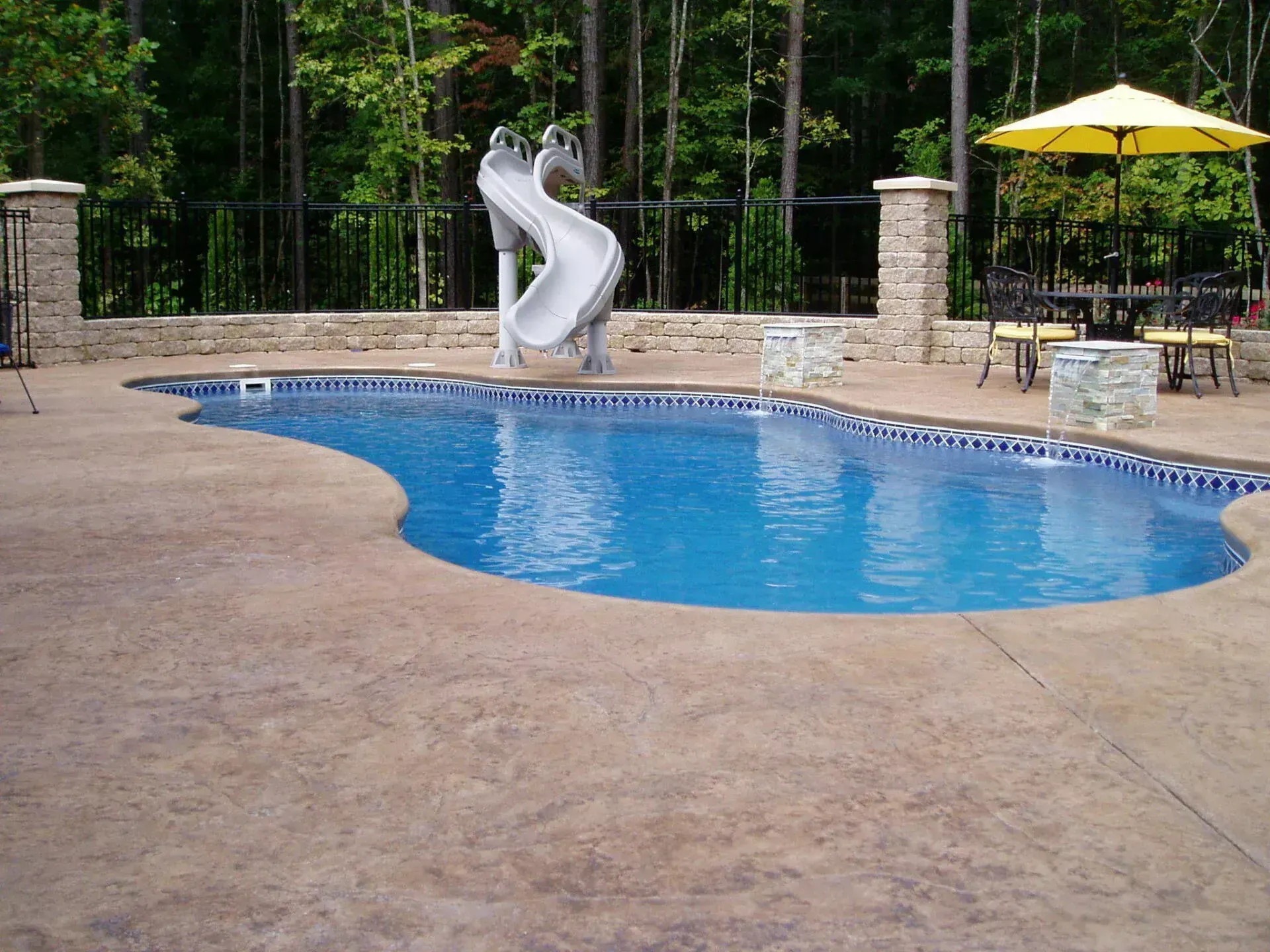 Swimming pool with a water slide, surrounded by a concrete patio, fence, and umbrella.