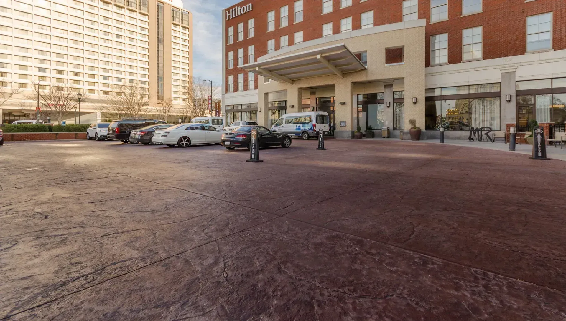 Hotel entrance with a brick-paved driveway. Cars are parked, and a large building is in the background.