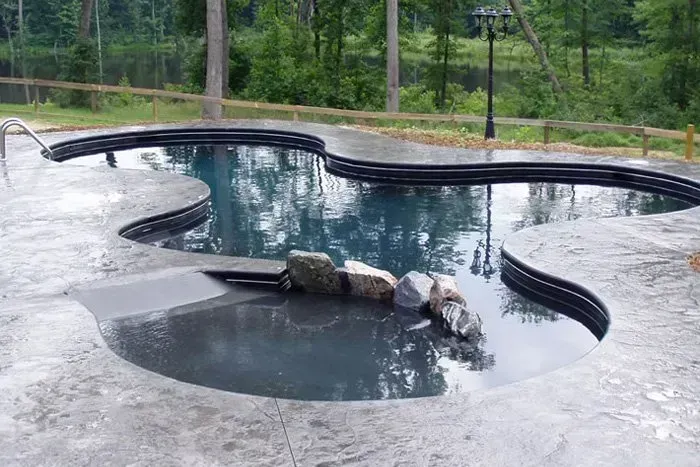 Freeform swimming pool with dark blue water and rock accents, surrounded by concrete and woods.