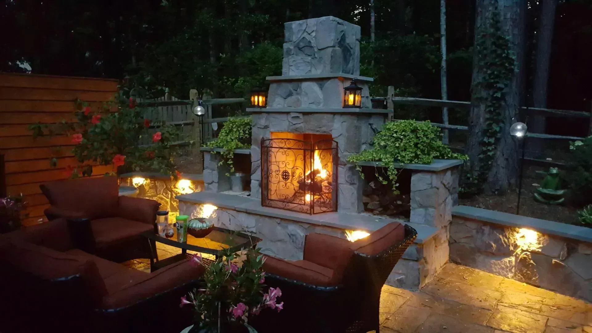 Outdoor stone fireplace with lit fire, surrounded by seating and lanterns.