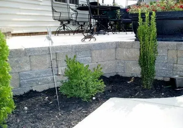 Stone retaining wall with bushes and plants, patio furniture visible in background.