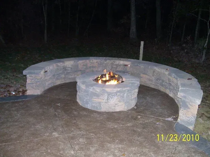 Stone fire pit with a burning fire, surrounded by a curved stone bench, set in an outdoor area at night.