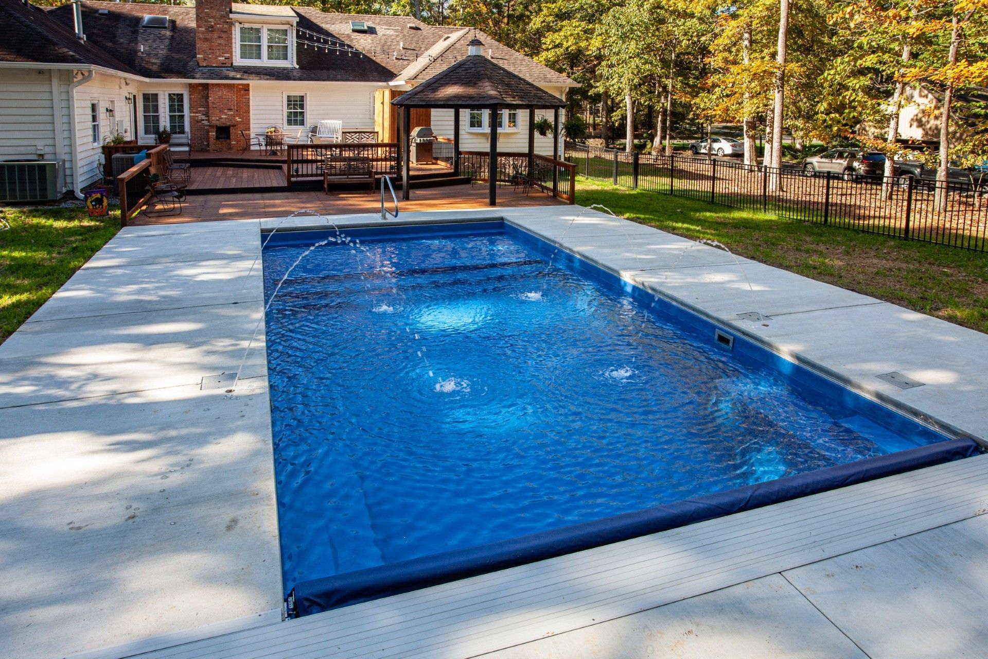 Rectangular blue swimming pool with fountains surrounded by concrete patio; backyard with house and gazebo visible.