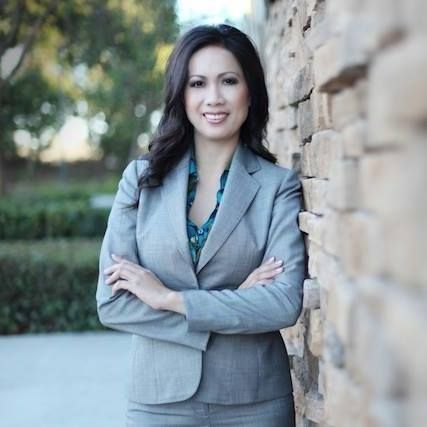 Woman in gray suit, arms crossed, leaning against a stone wall, smiling outdoors.