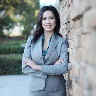 Woman in gray suit, arms crossed, leaning against a stone wall, smiling outdoors.